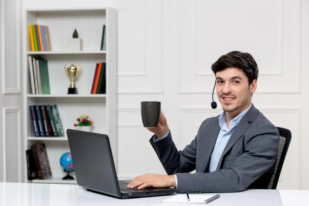 Man in a headset at a laptop, raising a mug